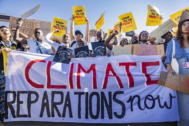 Under a blue sky, protesters hold a giant sign reading "Climate Reparations Now"