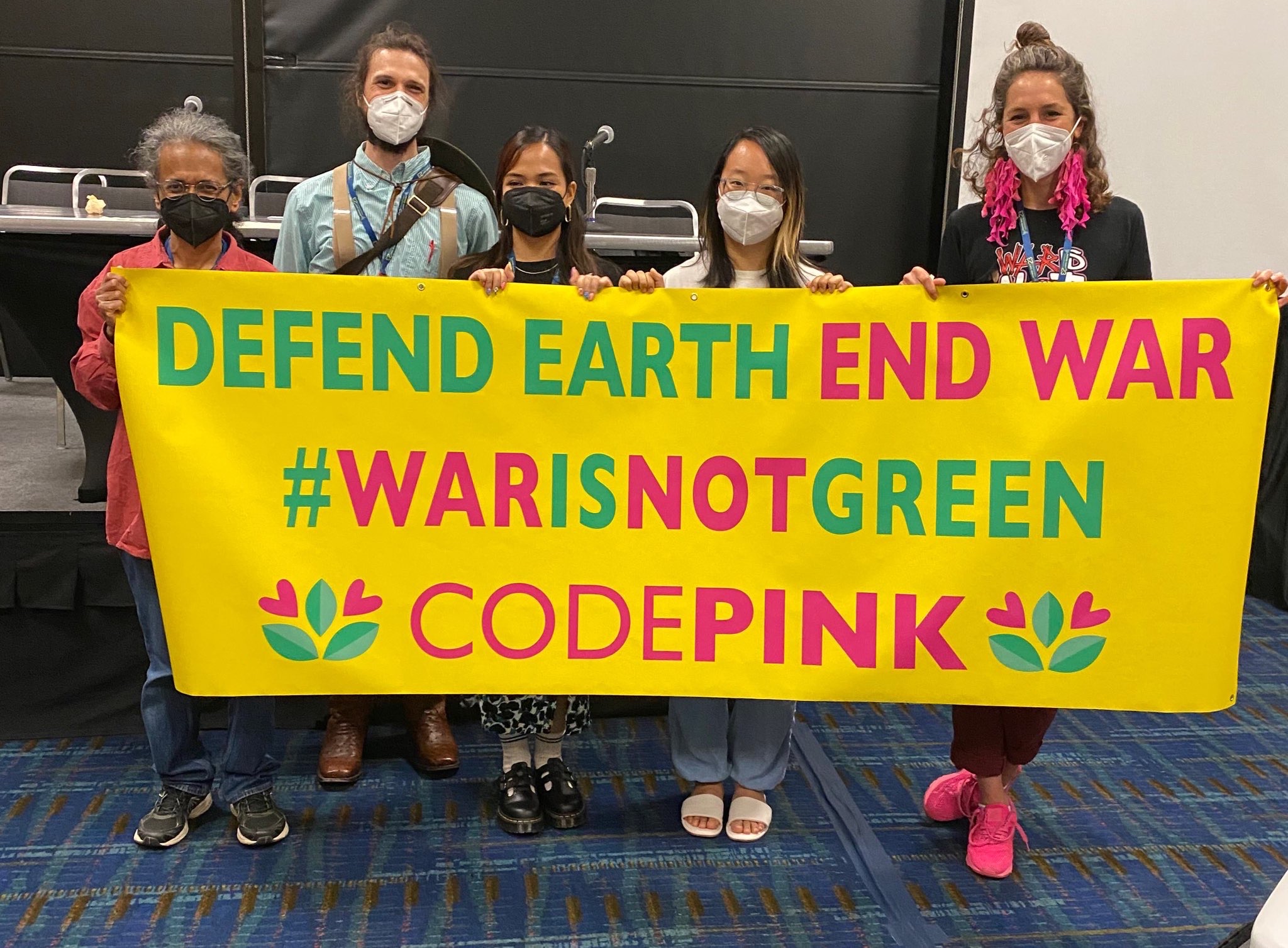 Five individuals from CODEPINK, Dissenters, Institute for Policy Studies, and Veterans for Peace hold up a banner saying "Defend Earth Not War #WarIsNotGreen CODEPINK"