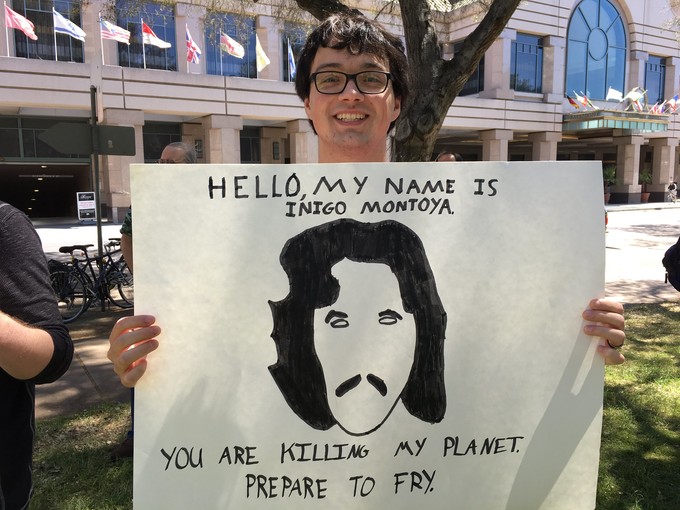 Climate protester in front of a large building holding a sign that reads, "Hello, my name is Inigo Montoya. You are killing my planet. Prepare to fry."