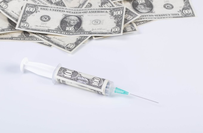 A hundred-dollar bill is rolled up inside a syringe, next to a pile of hundred-dollar bills