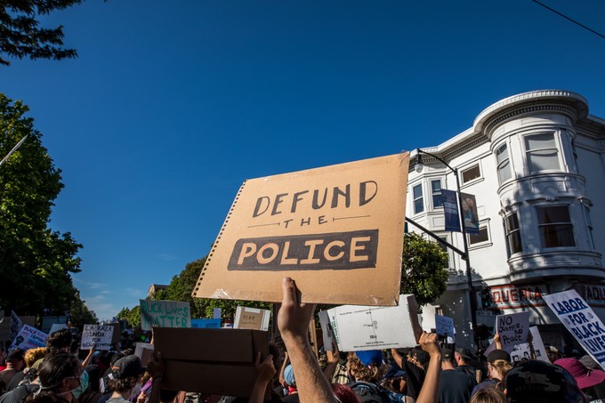 An arm rises above a protest crowd, holding a "Defund Police" sign against a blue sky and buildings